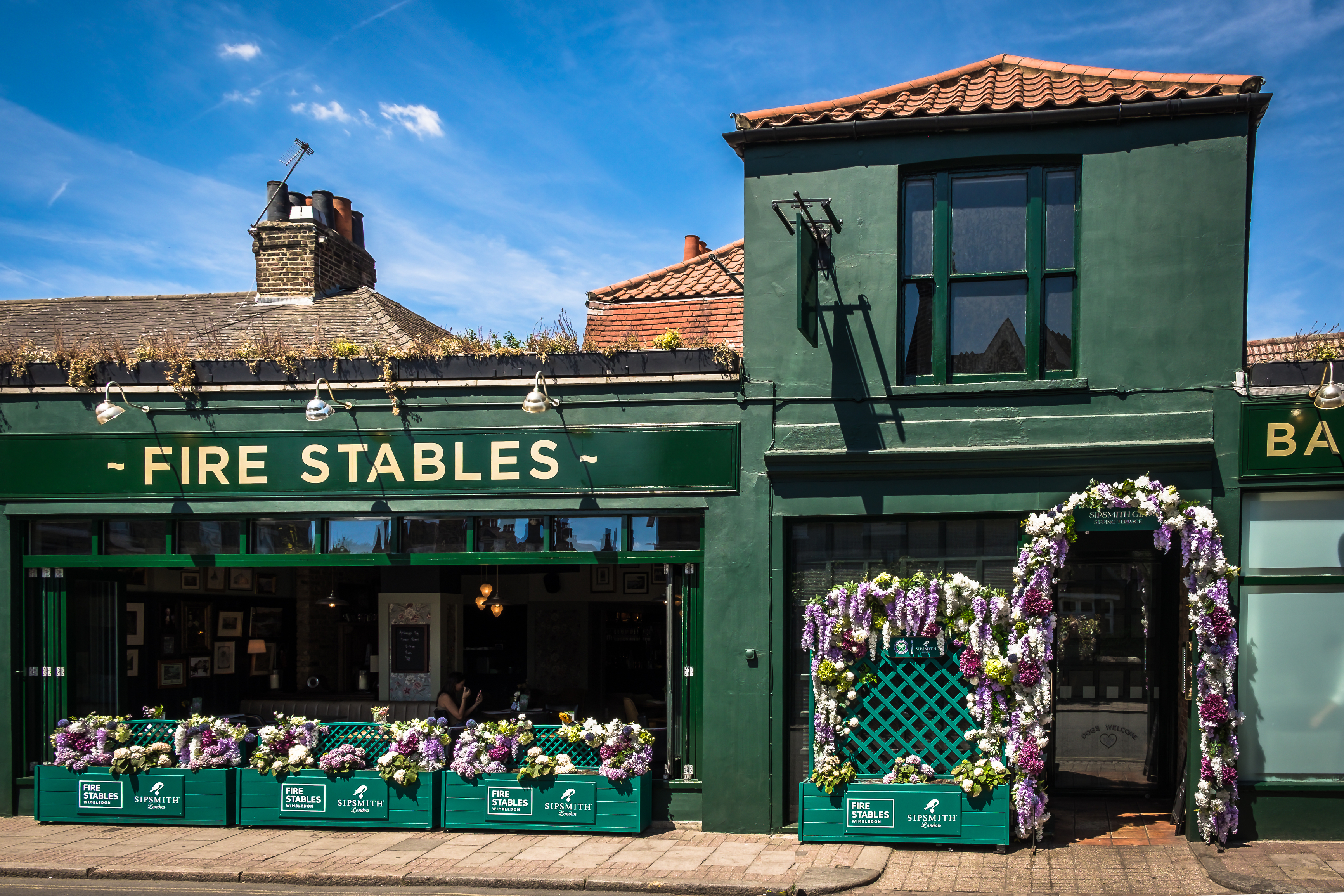 The green exterior of The Fire Stables in Wimbledon Village, decorated with flowers under a bright blue summer sky.